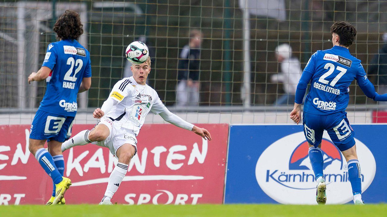A soccer player is kicking a ball on a soccer field. He is wearing a white jersey with logos. Behind him, there is a fence with a banner.