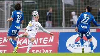 A soccer player is kicking a ball on a soccer field. He is wearing a white jersey with logos. Behind him, there is a fence with a banner.