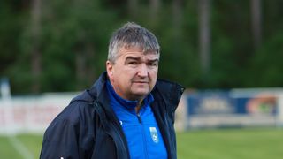 A man stands outdoors on a field wearing a blue shirt with a logo and a black jacket. The background is blurry with green trees.
