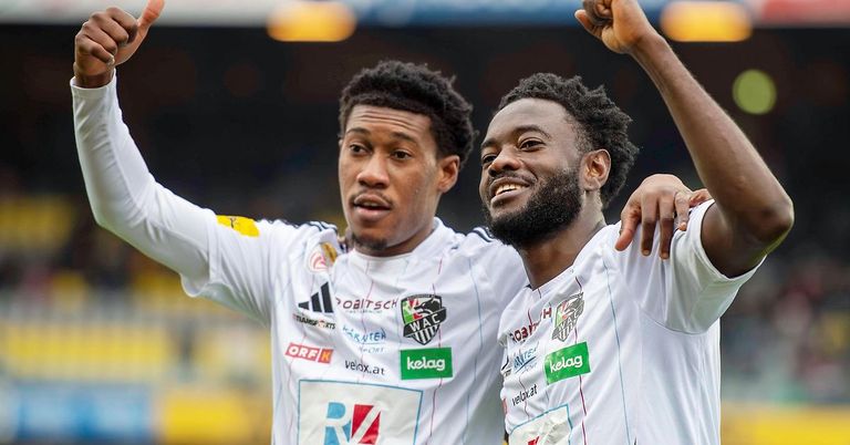 Two soccer players celebrate a goal, one with raised fists and the other smiling, both wearing white jerseys with various sponsors' logos.