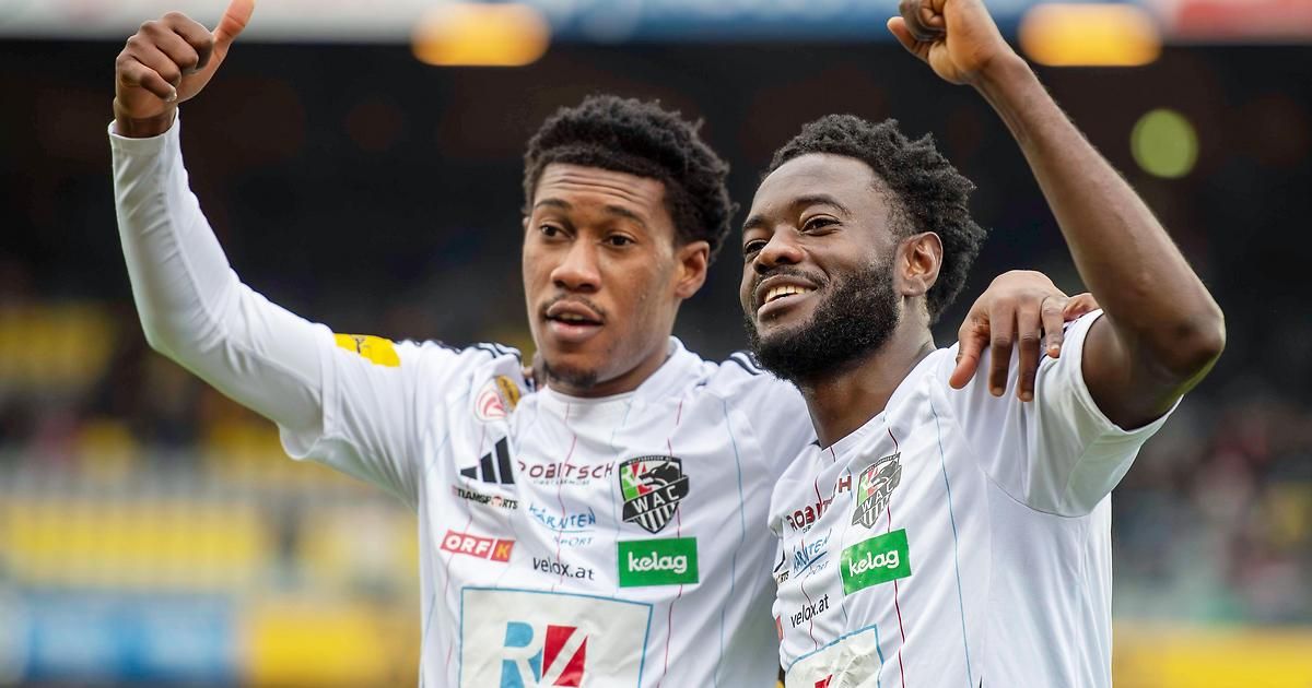 Two soccer players celebrate a goal, one with raised fists and the other smiling, both wearing white jerseys with various sponsors' logos.