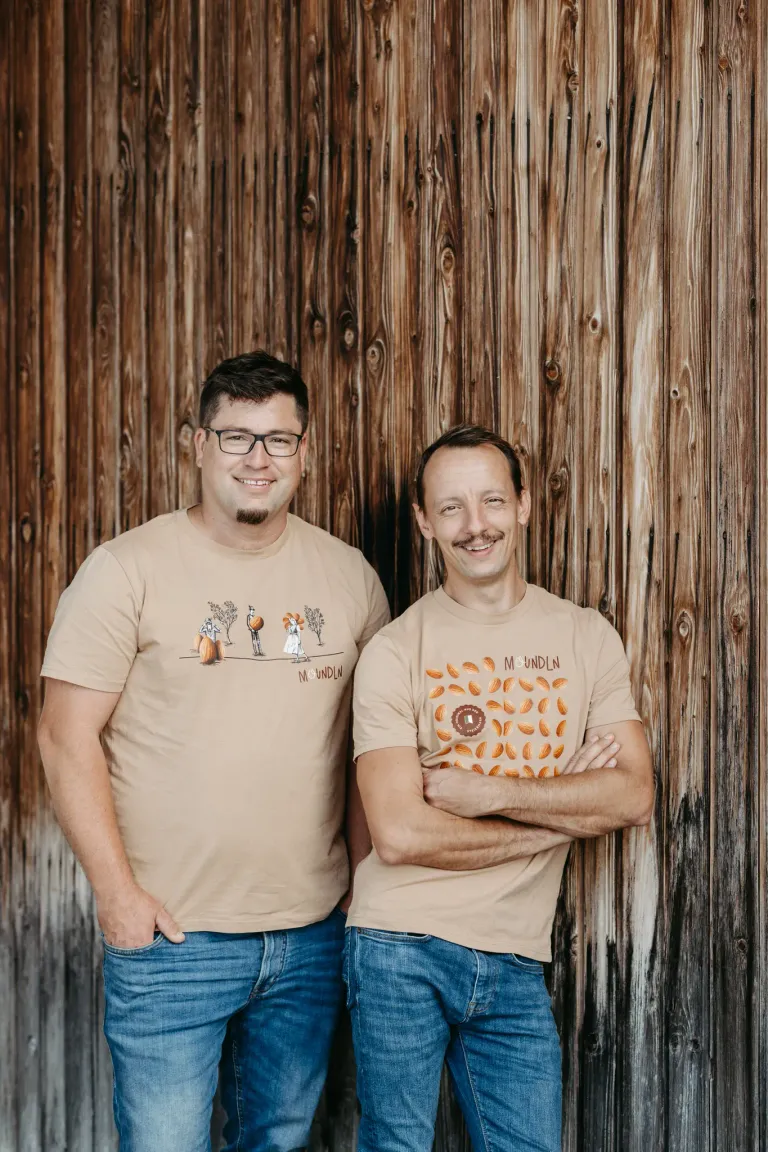 Two men stand in front of a wooden wall, both smiling and posing for a photo. The man on the left wears a shirt with a pumpkin illustration, while the man on the right wears a shirt with the text 'MOUNTLN'.