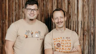 Two men stand in front of a wooden wall, both smiling and posing for a photo. The man on the left wears a shirt with a pumpkin illustration, while the man on the right wears a shirt with the text 'MOUNTLN'.
