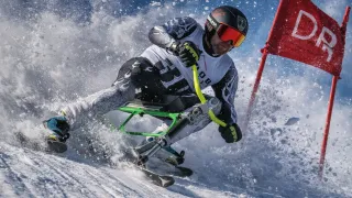 A skier wearing a helmet and goggles is riding a specialized ski board down a snowy slope, creating a spray of snow.