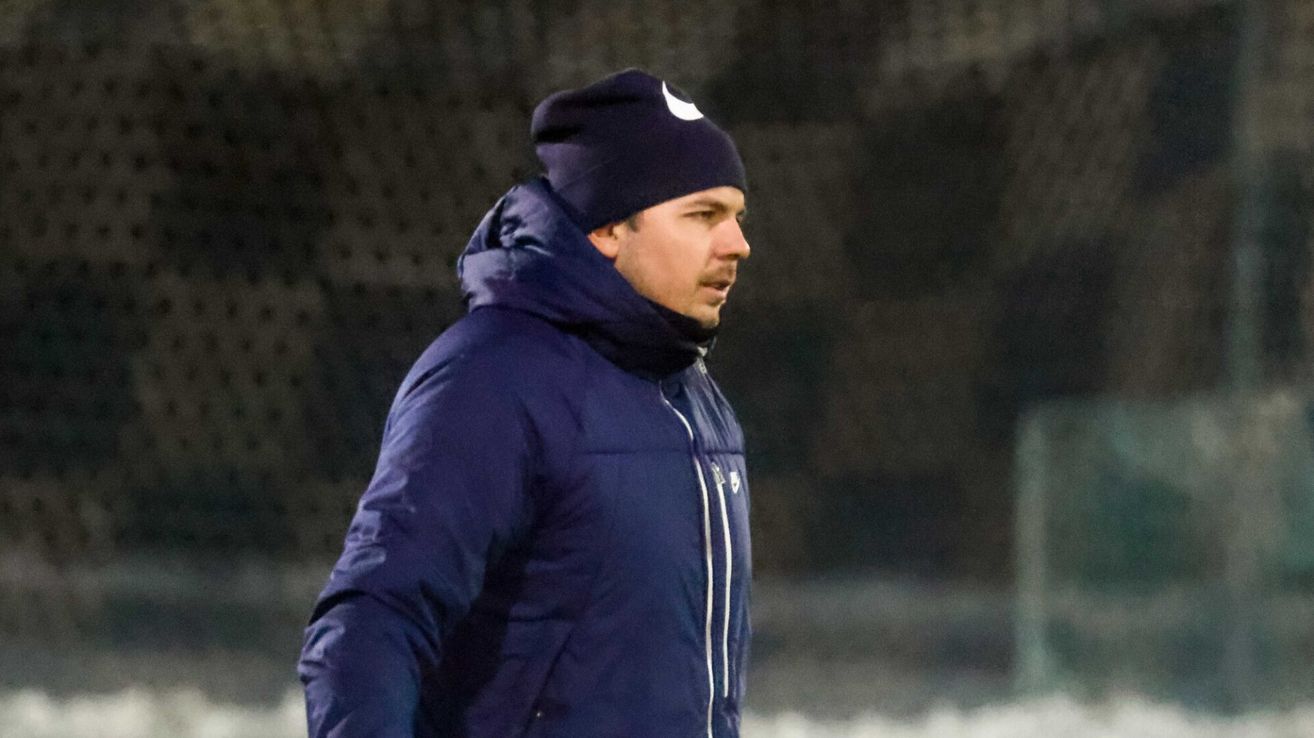 A man in a navy blue jacket and beanie walks on a field, looking to the right. The background is blurred, indicating he is in motion.
