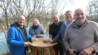 Five people, including a woman, are standing around a small wooden table, smiling for a picture. They are outdoors with trees in the background.
