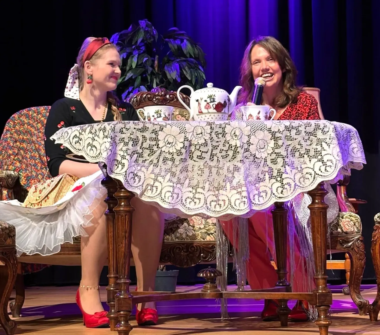Two women are seated on stage, one in a traditional dress, the other with a microphone, smiling. They sit at a table with a teapot and cups. The stage has a backdrop and a plant.