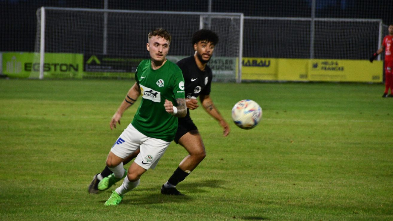 Two soccer players in action, one in green and one in black, running towards a ball. They are on a field with a goal net in the background.