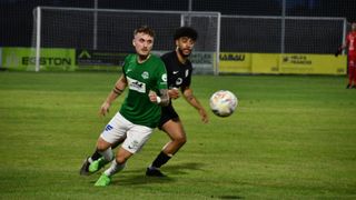 Two soccer players in action, one in green and one in black, running towards a ball. They are on a field with a goal net in the background.