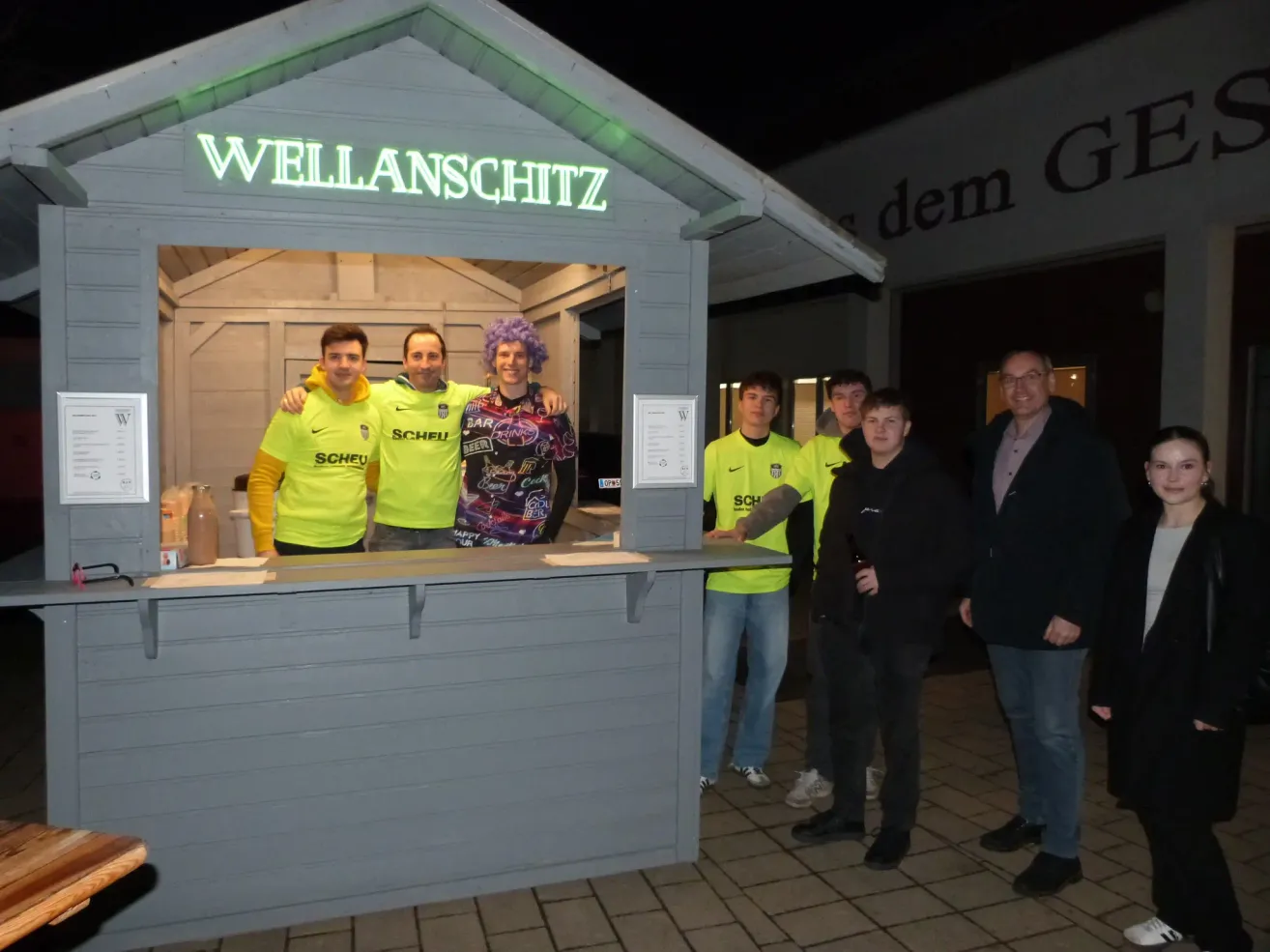 A group of men stand in front of a booth labeled Wellanschitz. They wear neon yellow jerseys with Scheu on them. Behind them, another man stands next to a sign.