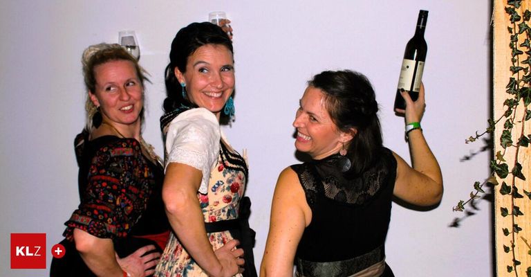 Three women standing close together, smiling and holding drinks. The woman in the middle holds a wine bottle. They seem to be posing for a photo.