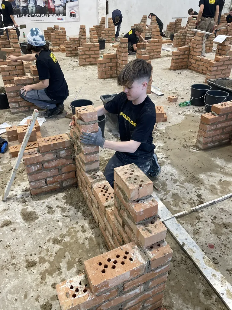 Zwei Personen in schwarzen Shirts und Handschuhen arbeiten an einer Ziegelmauer, eine kniet und eine sitzt. Das Gebiet ist von anderen Ziegelstrukturen und Eimern umgeben.