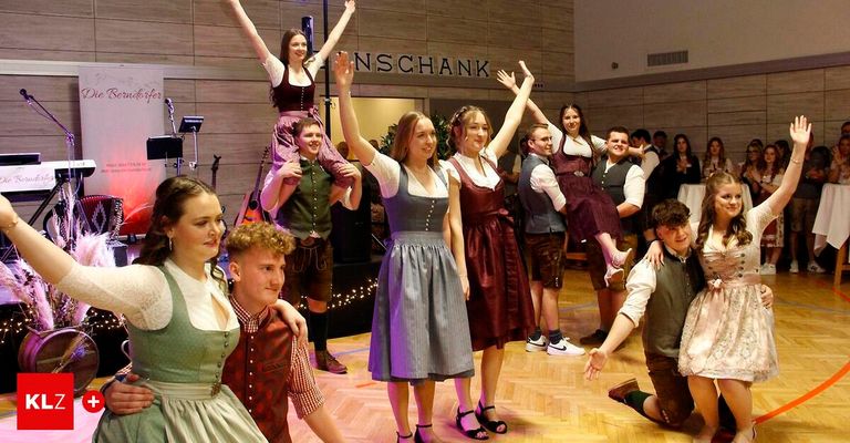 Mehrere Menschen in traditioneller deutscher Kleidung tanzen auf einem Holzboden. Eine Frau steht auf den Schultern zweier Männer. Musikinstrumente sind auf der Bühne. Die Wand hinter ihnen hat das Wort 'Inschank' darauf.