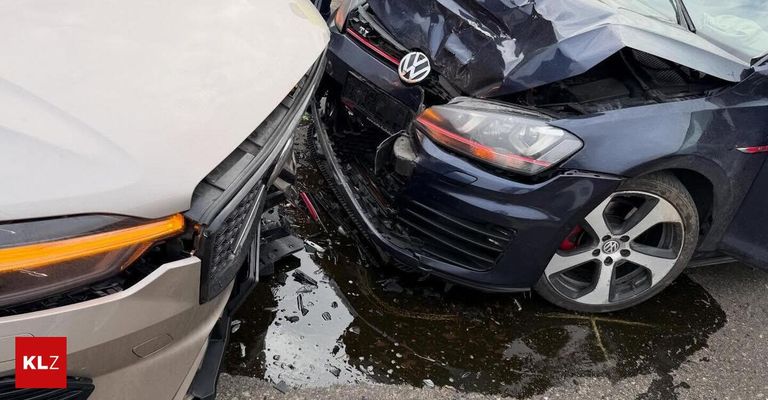 Ein weißer Wagen und ein blauer Volkswagen mit beschädigten Fronten stehen in einer Pfütze auf einer nassen Straße. Der Stoßfänger des Volkswagens ist verbeugt und die Frontgrille ist beschädigt.
