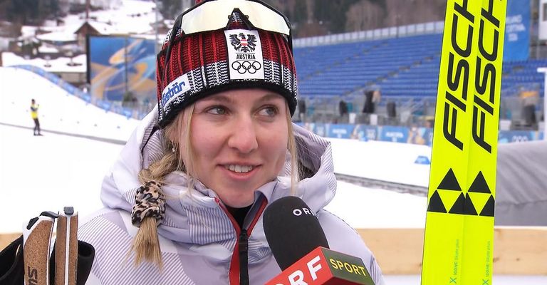 A woman with a snowy Austrian beanie speaks into a microphone. She is smiling and appears to be at a ski event.
