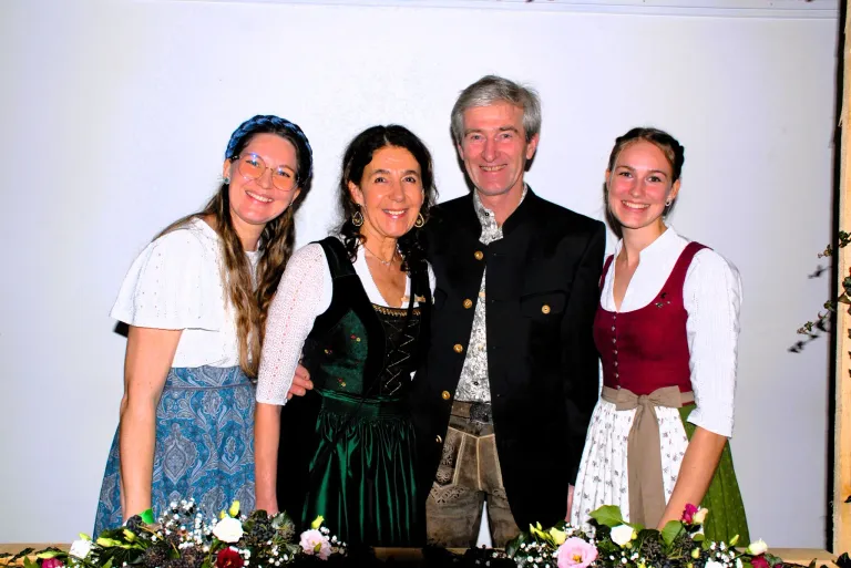 Five people stand together for a photograph, smiling. Three women and one man. All are dressed in traditional Bavarian attire. The woman in the middle wears a green dress and necklace. The man wears a black coat.