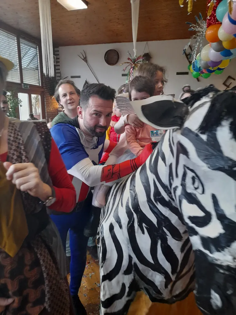 A man holds a child up to a zebra statue while a woman and others look on. The setting appears to be indoors with balloons and decorations in the background.