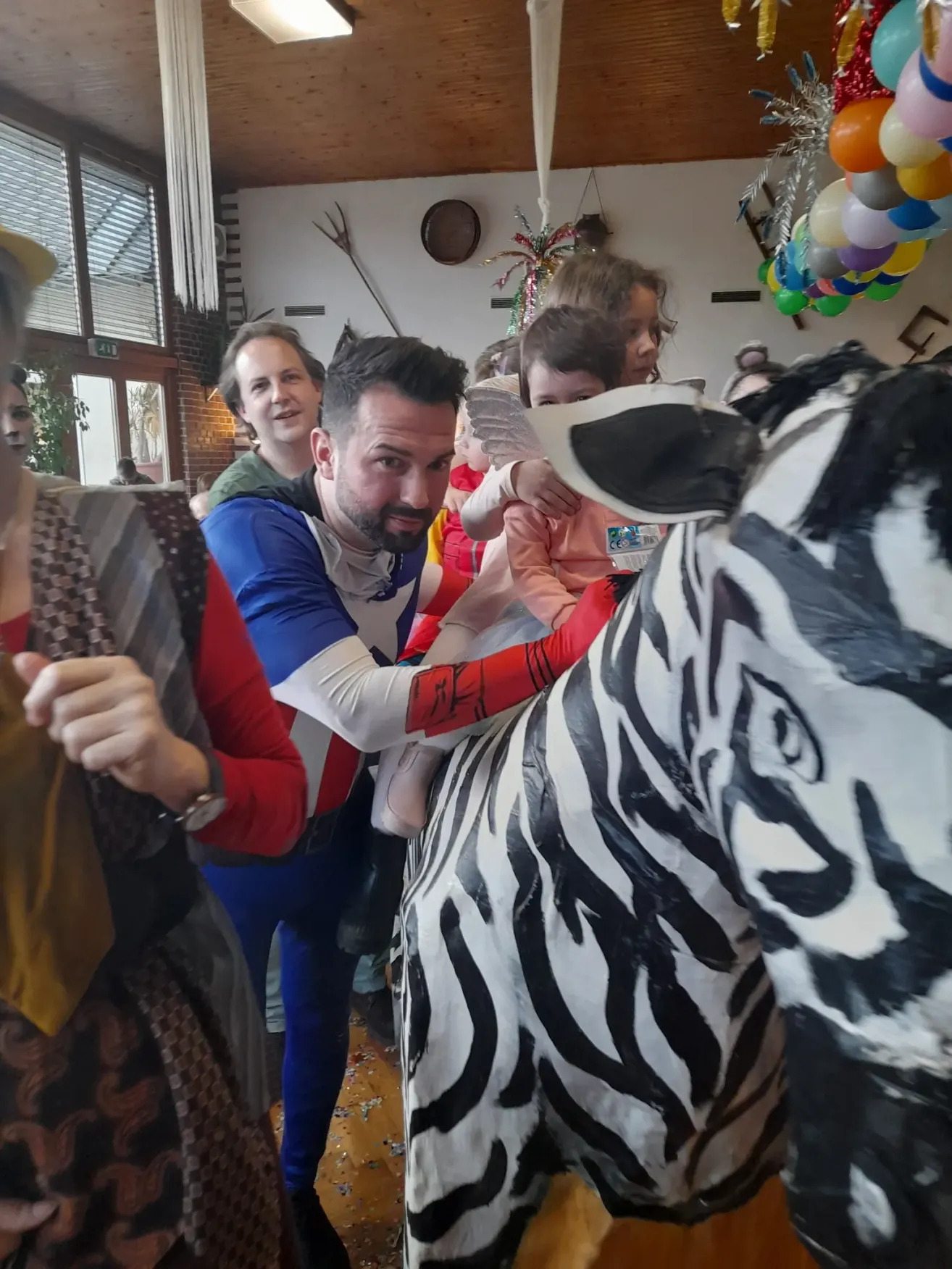 A man holds a child up to a zebra statue while a woman and others look on. The setting appears to be indoors with balloons and decorations in the background.