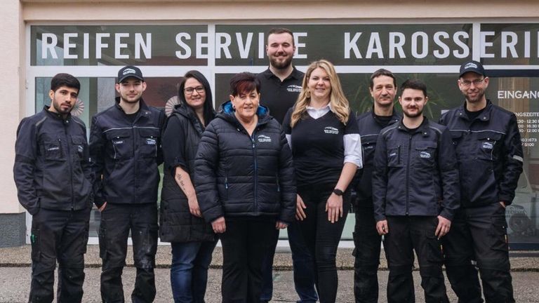 Eine Gruppe von Menschen in dunklen Jacken und Hosen steht vor einem Gebäude mit einem Schild, das 'EN SERVICE KARO' besagt.