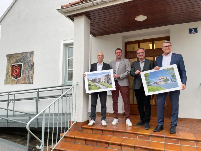 Vier Männer stehen auf einer Treppe und halten Baupläne in der Hand. Sie sind in formeller Kleidung gekleidet. Der Mann auf der linken Seite trägt eine Brille und Turnschuhe. Das Gebäude hat ein Holzdach, eine weiße Wand und eine Holztür.