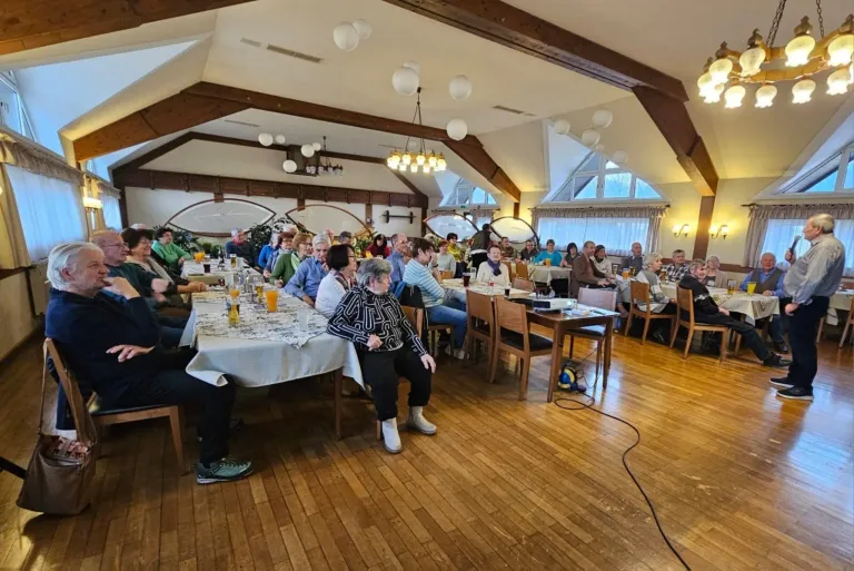 A dining hall with wooden floors and tables covered in white cloth. People are seated around the tables, some are drinking, and some are engaged in conversation. There are lamps hanging from the ceiling.