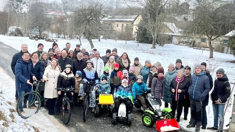 Eine Gruppe von Menschen in Winterkleidung steht und sitzt auf Fahrrädern und kleinen Fahrzeugen in einem verschneiten Bereich, mit Gebäuden im Hintergrund.
