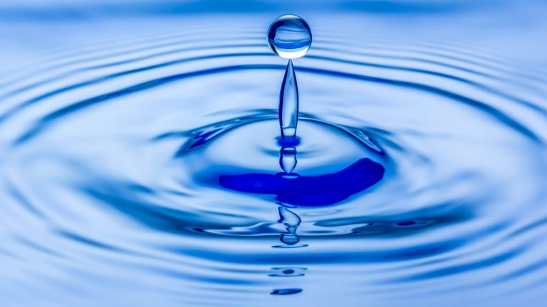 A water droplet is falling into a blue water surface creating ripples. The droplet is in the center of the image.