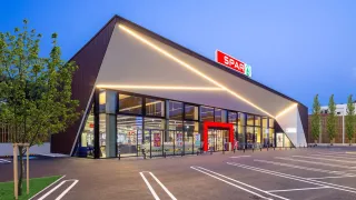 The front of a Spar store, illuminated at night, with a large glass facade, a red entrance, and a sign above the entrance.