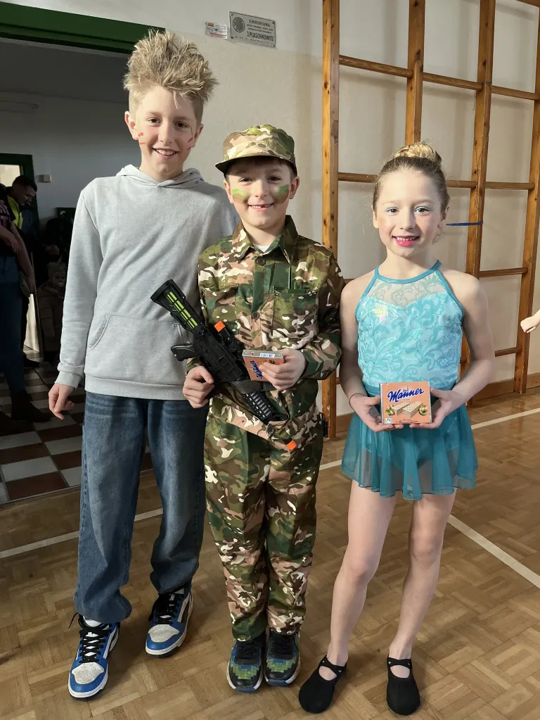 Three children are standing together in a room. The boy in the middle is dressed in a camouflage uniform and holding a toy gun. The boy on the left is wearing a gray hoodie. The girl on the right is holding a box of cookies.