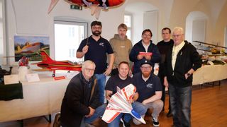 A group of men pose for a photo in a room with models and a banner, smiling and holding a paper airplane.