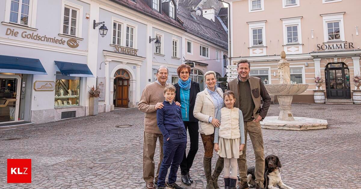 A family of five, including two adults and three children, poses for a photo in front of an old town square with cobblestones, a fountain, and buildings.