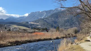 A scenic view of a river flowing through a valley with mountains and houses. The mountains are covered with snow.