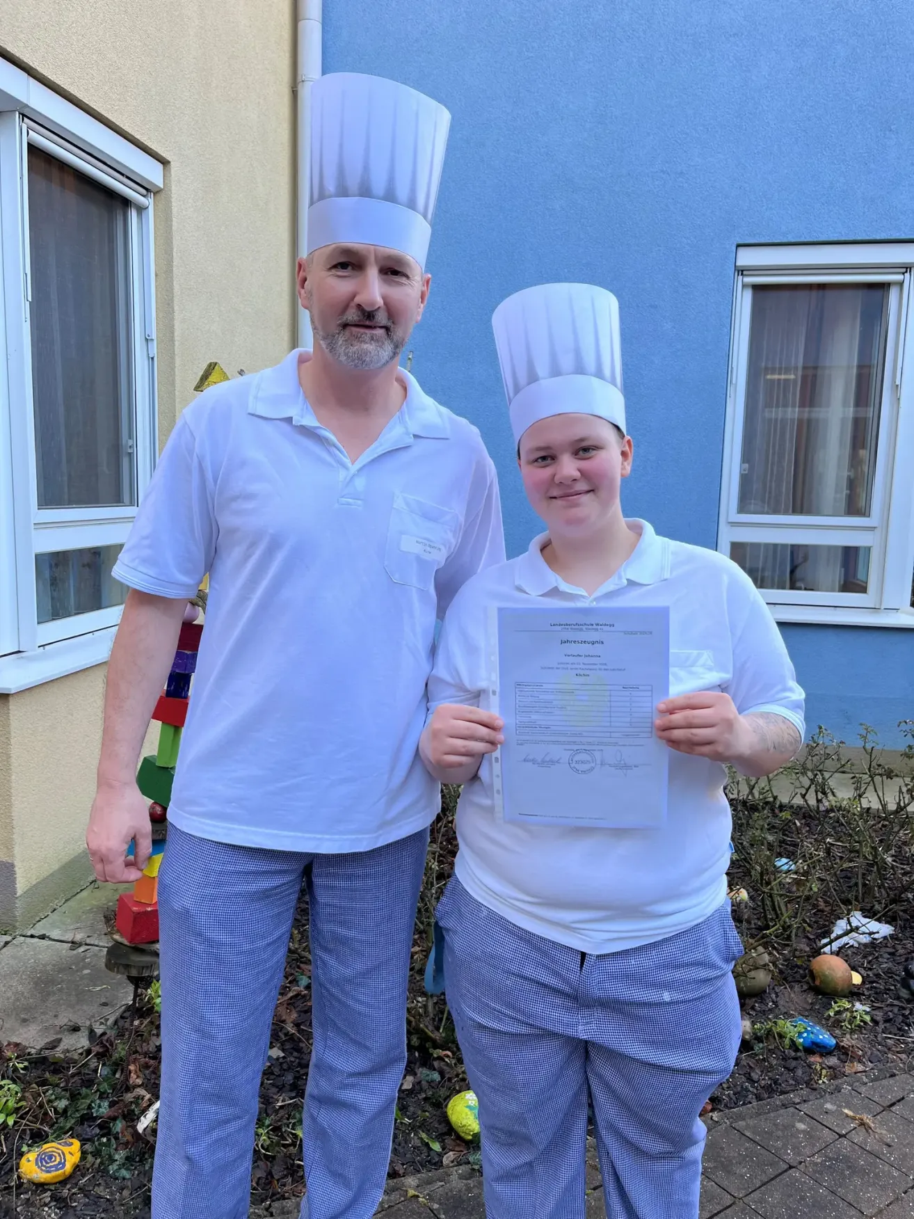 Two individuals in chef hats stand in front of a building, one holding a certificate. The man has a beard, and the woman smiles. Behind them, a window and a door are visible, with a small structure and a pile of toys on the ground.