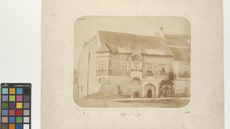 A vintage photograph shows an old, weathered building with a gabled roof, several windows, and a grand entrance with a wooden door. Two people stand near the entrance.