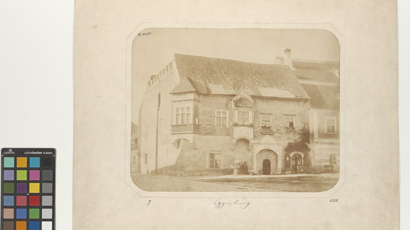 A vintage photograph shows an old, weathered building with a gabled roof, several windows, and a grand entrance with a wooden door. Two people stand near the entrance.