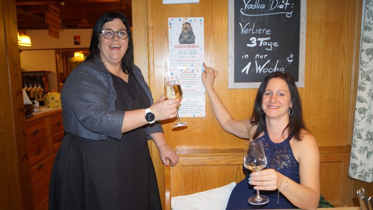 Two women celebrate with wine glasses, standing next to a wooden wall. One wears glasses and a watch, the other a blue dress. Behind them is a poster and a chalkboard with writing.