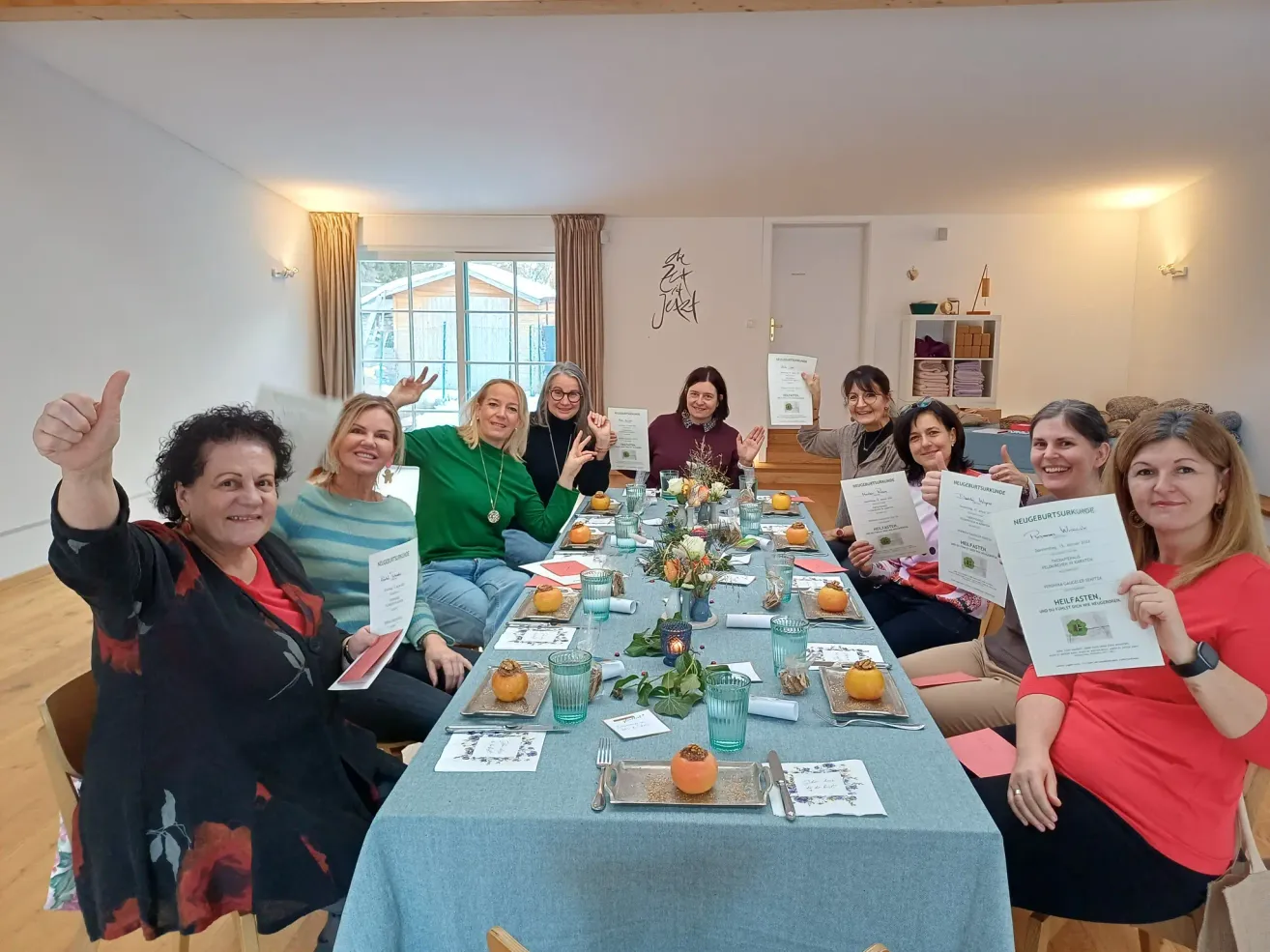 A group of women sit at a table holding certificates. They are smiling and posing for a photo. The table is covered with a blue cloth and has food and flowers on it. Behind them, there is a wall with a painting.