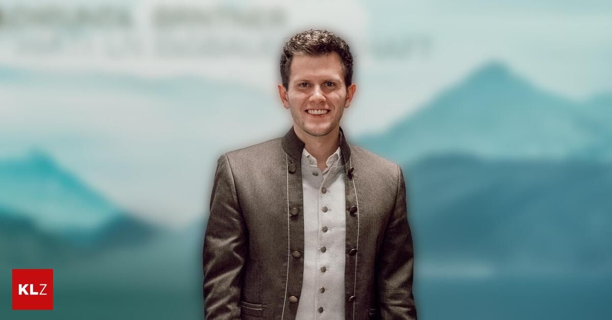 Ein Mann mit kurzen braunen Haaren lächelt in die Kamera. Er trägt einen grauen Mantel mit Knöpfen über einem weißen Hemd. Der Hintergrund ist unscharf, mit blauen Bergen.