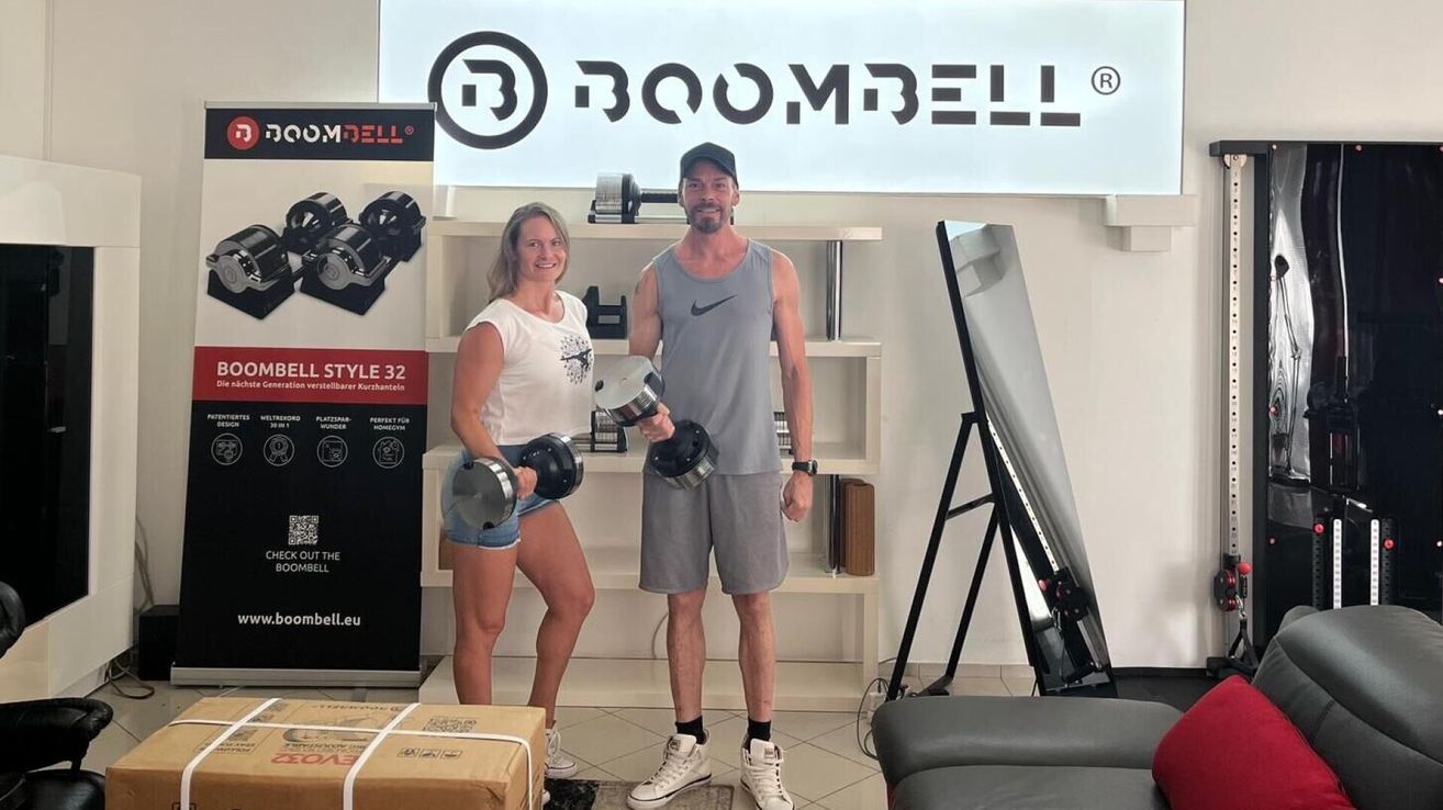 A man and a woman stand in a gym, holding dumbbells. Behind them, shelves display various weights and a sign with the text 'Boombell'. They both smile and appear to be posing for a photo.