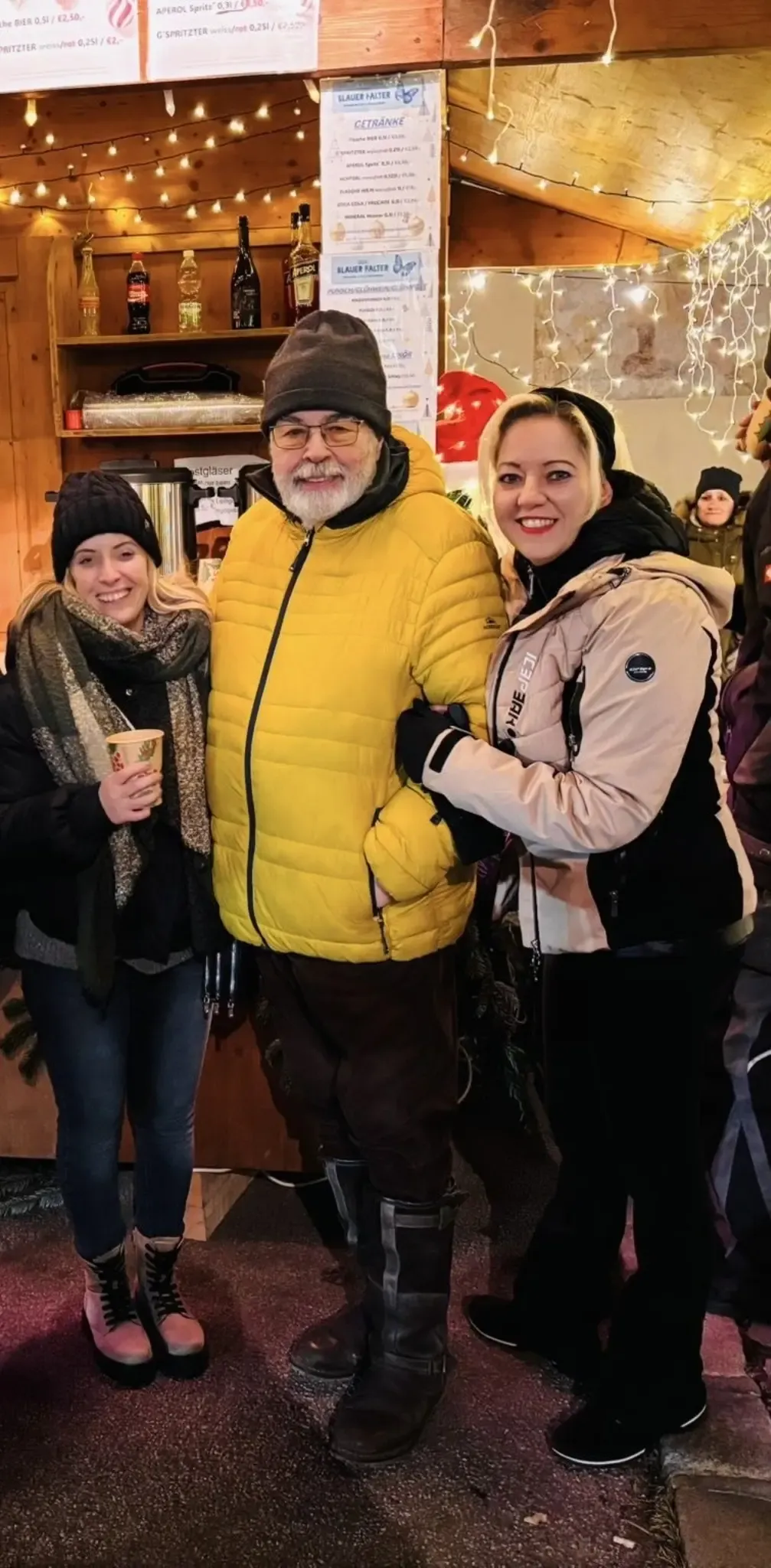 Three people are standing together and smiling. The woman on the left is holding a cup, while the man in the middle is wearing a yellow jacket. The woman on the right is wearing a beige jacket. Behind them is a wooden shelf with coffee machines and other items.