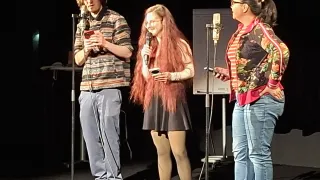 Three people on a stage with microphones. A man on the left with a phone, a woman in the middle with long hair and a microphone, and a woman on the right with glasses and a microphone. Behind them is a whiteboard.