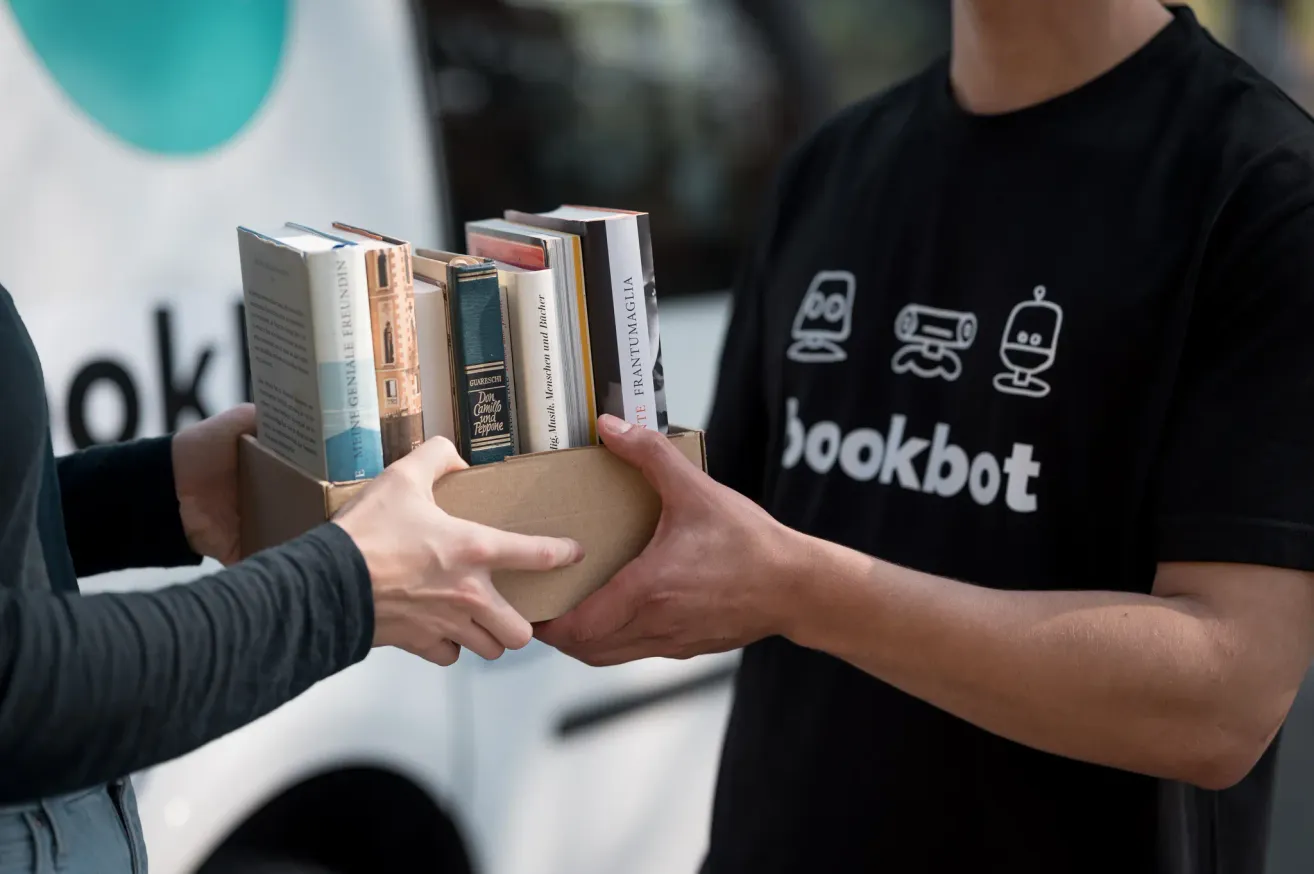 Two hands are holding a cardboard box filled with books. The person in a black t-shirt is wearing a Bookbot shirt. The person in a long-sleeve shirt is holding the box.