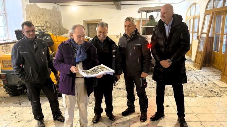 Vier Männer in Winterkleidung stehen in einem Raum. Der Mann auf der linken Seite hält ein Papier, während die anderen ihn ansehen. Hinter ihnen ist eine gelbe Maschine zu sehen.