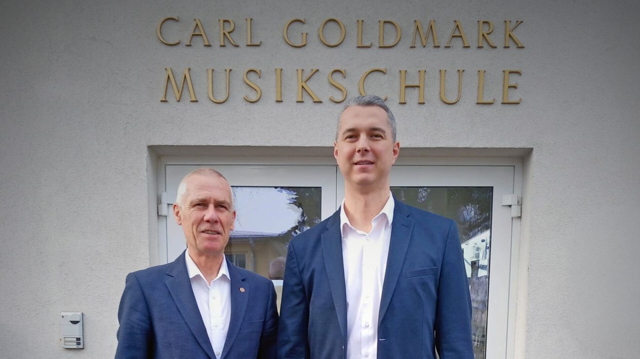 Two men in suits stand in front of the Carl Goldmar Musikschul building, smiling at the camera.