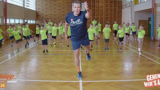 A man leads a group of young children in a gymnasium, all dressed in green shirts and shorts, doing jumping jacks.