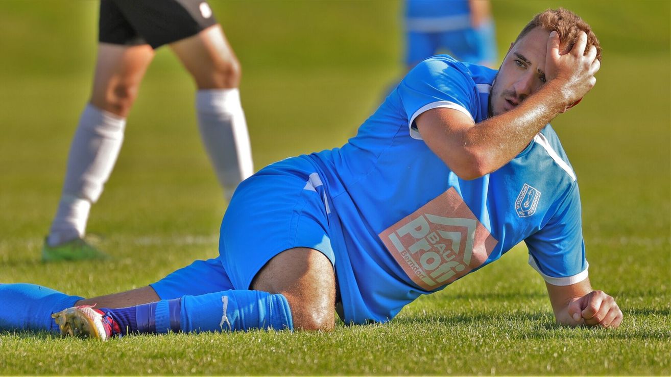 A soccer player in a blue jersey lies on the grass with his head on his arm. Behind him, another player stands in blue shorts.