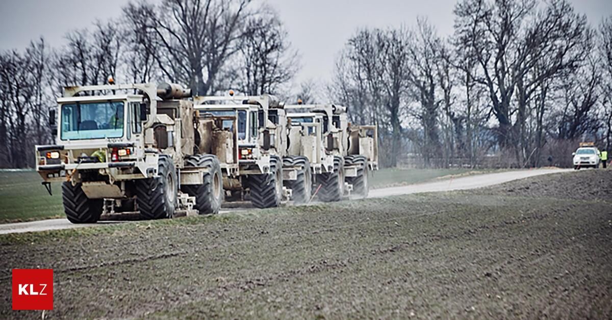 A line of military trucks drives on a dirt road in a field, surrounded by bare trees.
