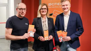 Three people stand together in front of a banner with text reading 'Meine Stadt, Meine App' and 'CITIES schon'. The man on the left holds an orange booklet, the woman in the middle holds an orange booklet, and the man on the right holds two orange booklets.