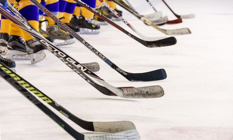 Multiple hockey sticks lined up on ice, showing different brands and designs. Players in yellow, blue, and black jerseys with skates are standing behind the sticks.