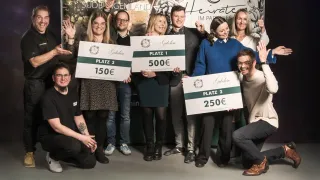 A group of people pose with signs displaying awards, including Platz 1 with 500€, Platz 2 with 250€, and Platz 3 with 150€. They stand in front of a backdrop with floral and text design.